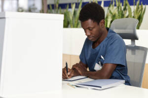 Young African American male doctor or administrator of clinics making notes in medical document in clipboard while sitting by counter